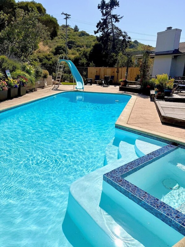 A clear blue pool with a slide, surrounded by a wooden deck and potted plants.