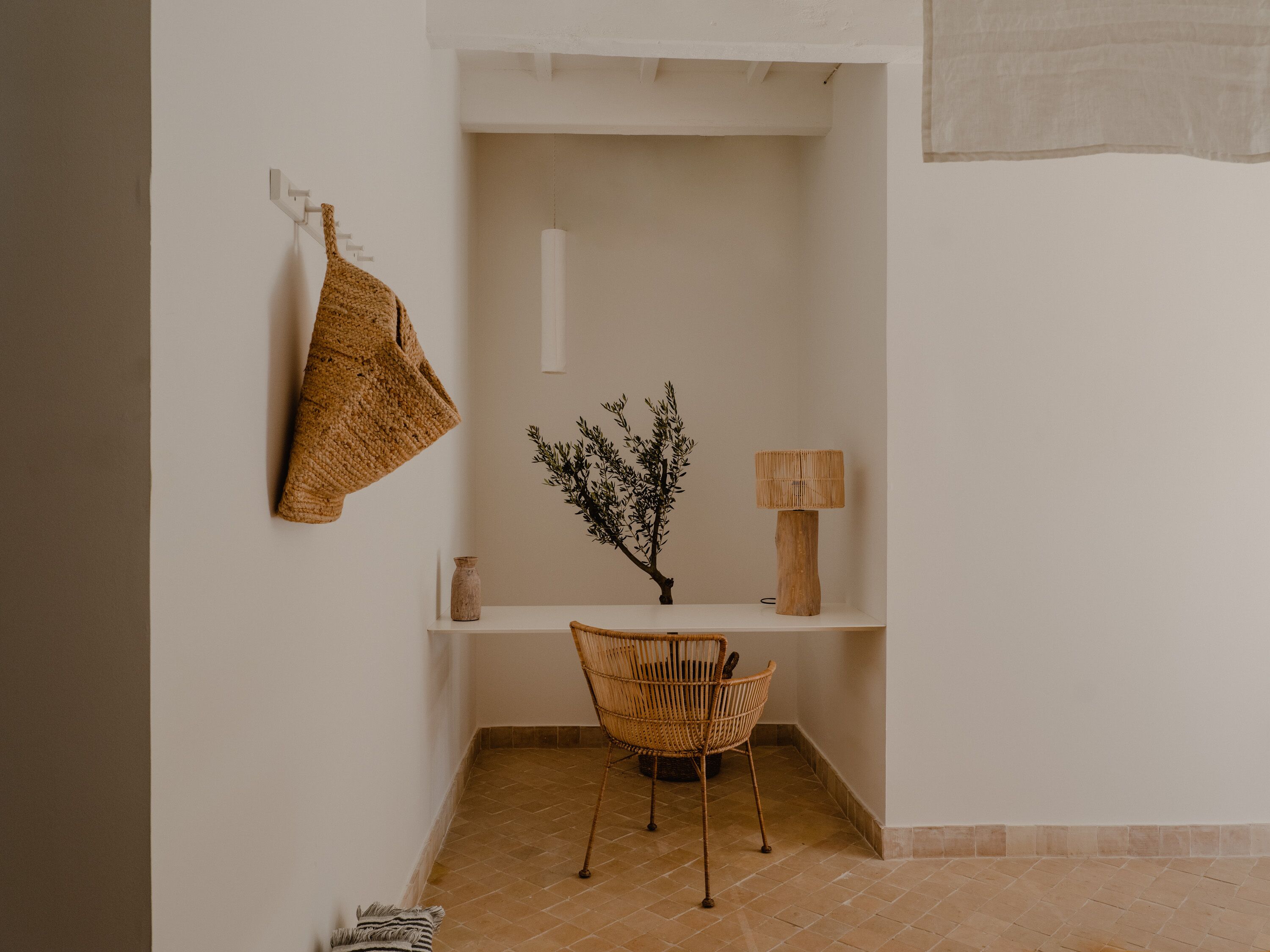 A corner room with a white wall, a woven chair, a small table with a lamp, and a hanging towel.
