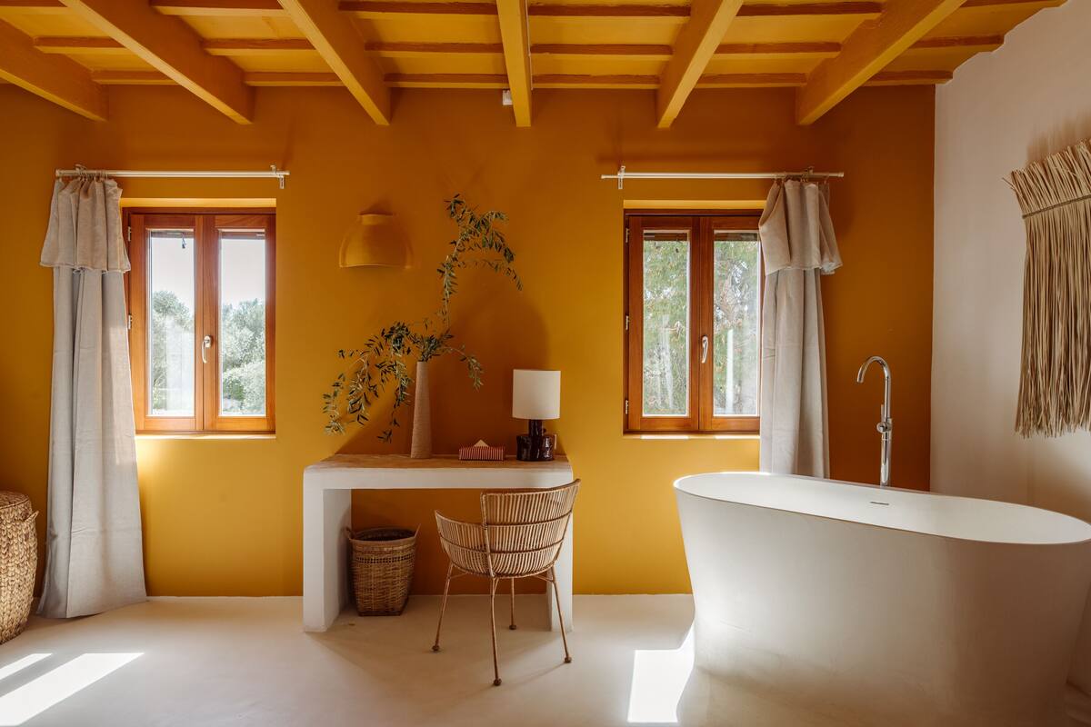 A bathroom with a bathtub, a desk, and a chair. There are two windows with curtains, a plant on the desk, and a lamp.