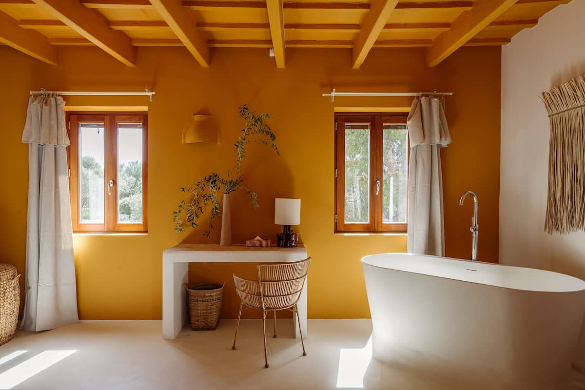 A bathroom with a bathtub, a desk, and a chair. There are two windows with curtains, a plant on the desk, and a lamp.