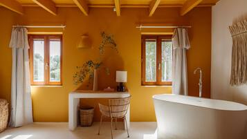 Un baño con bañera, un escritorio y una silla. Hay dos ventanas con cortinas, una planta en el escritorio y una lámpara.