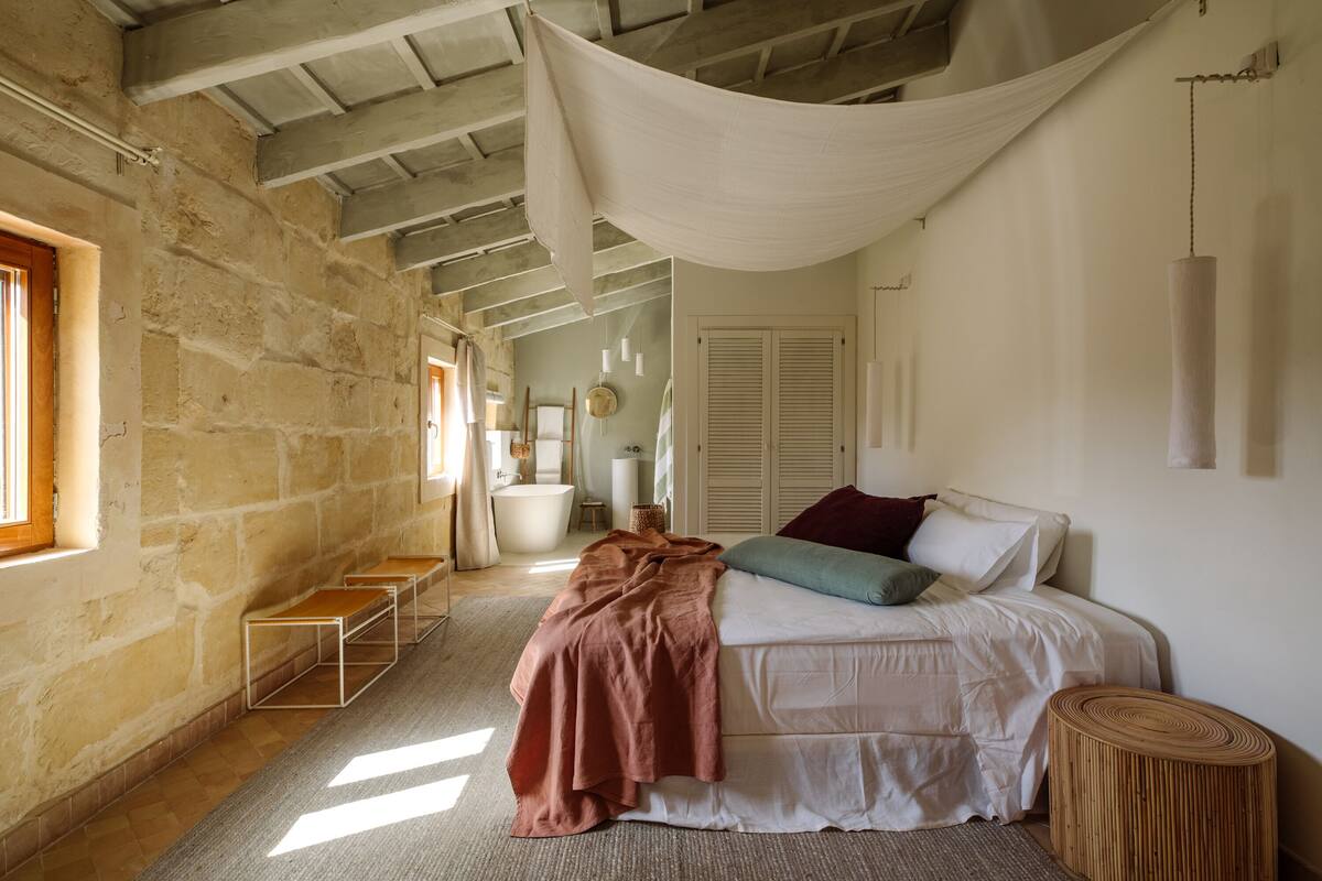 A bedroom with a bed, a window, a wooden bench, and a hanging light fixture.
