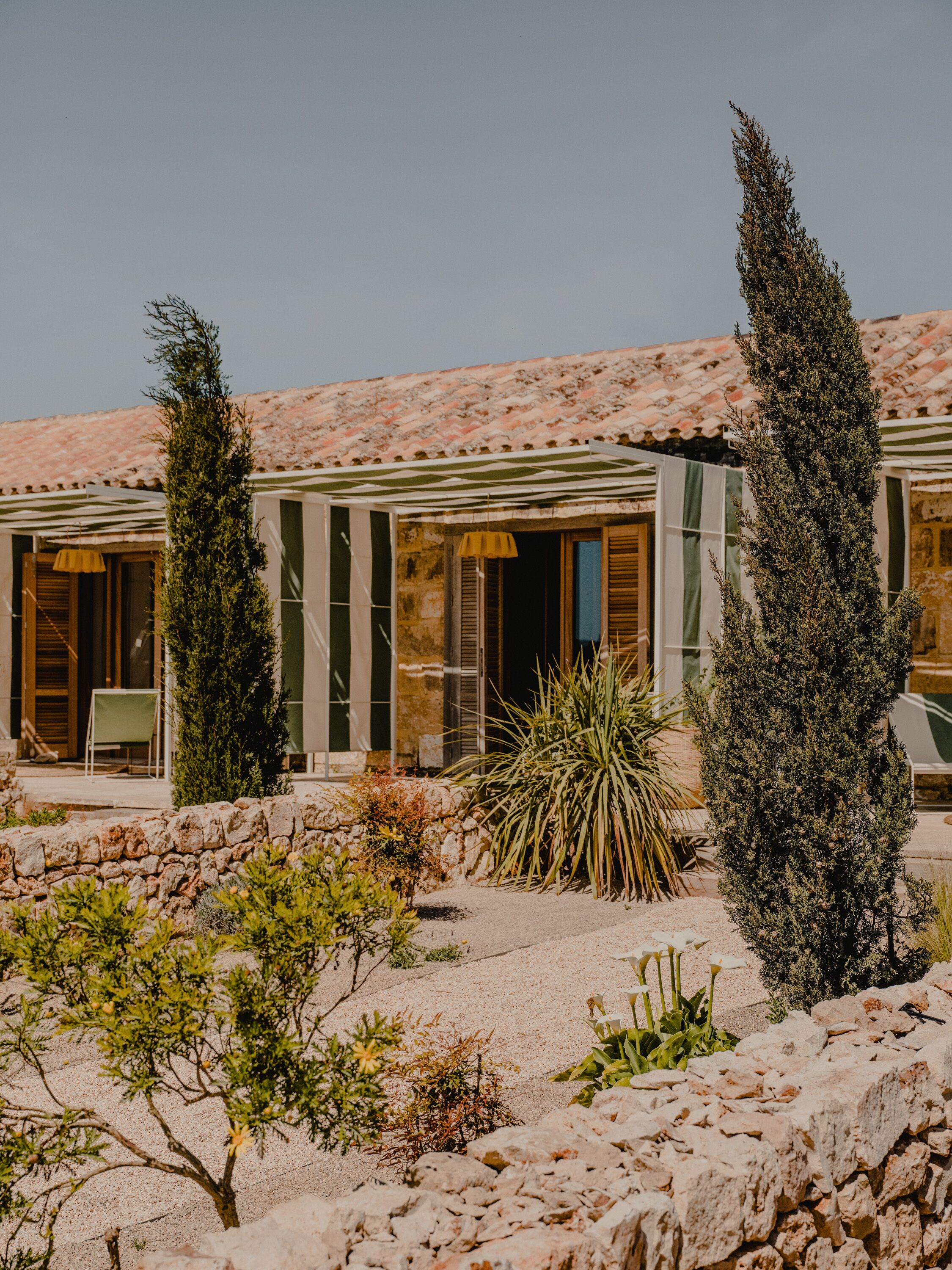 A traditional Mediterranean-style building with a tiled roof, stone walls, and a garden with various plants and a stone wall.