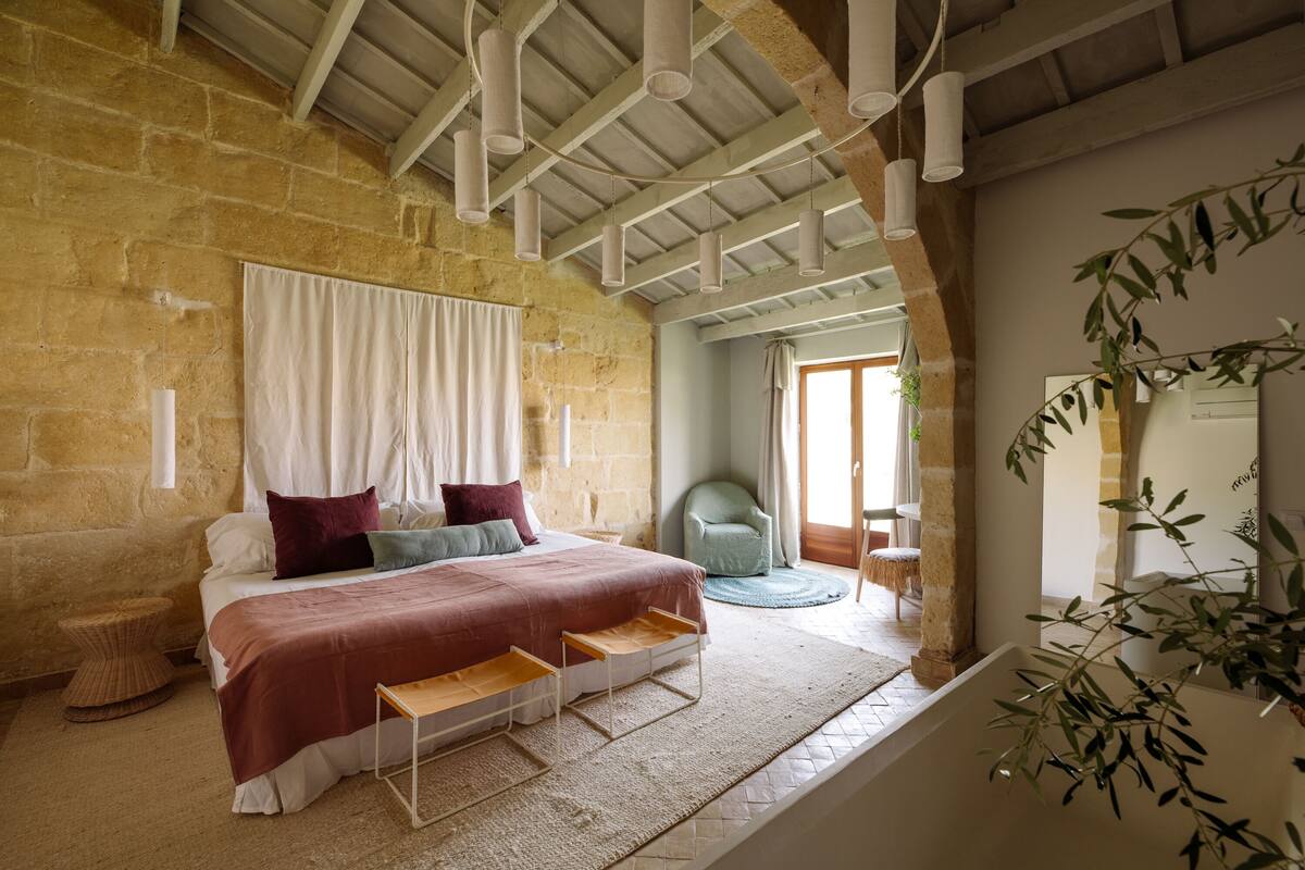 A bedroom with a large bed, a small table, a chair, and a window with curtains.