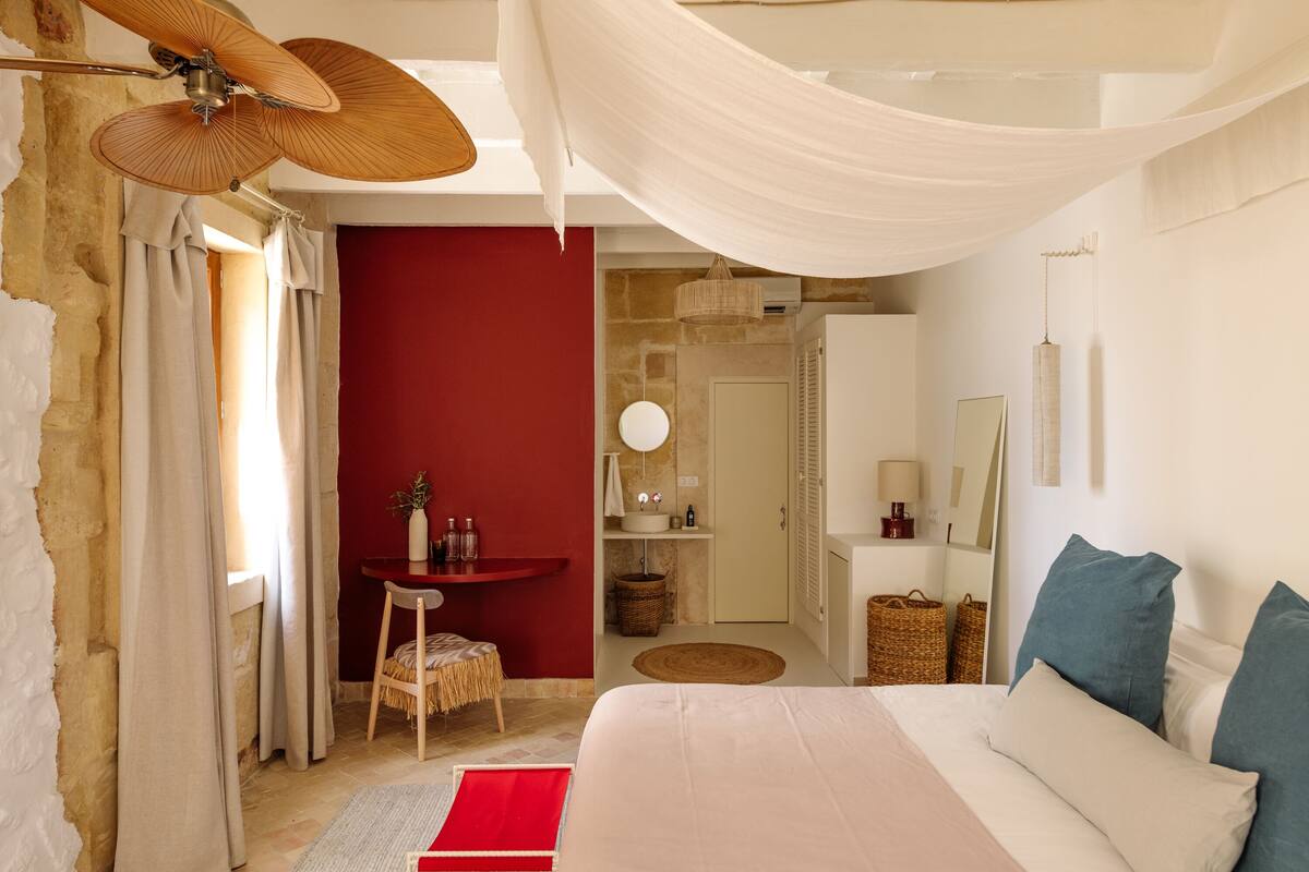 A bedroom with a bed, a desk, a chair, a red wall, a mirror, and a hanging light fixture.