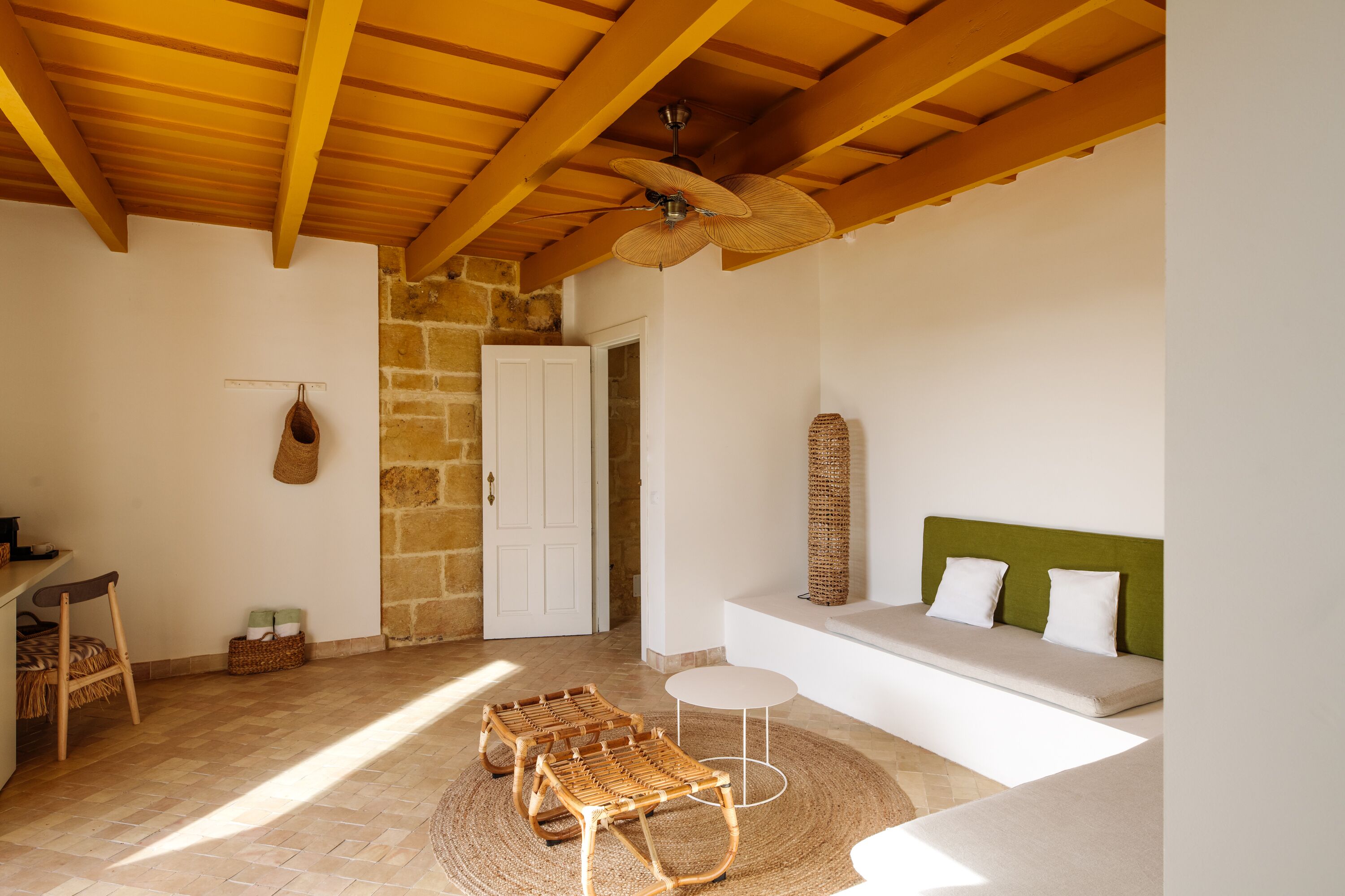 Une pièce avec un plafond en bois, un mur en pierre, une porte blanche, un canapé vert avec des coussins blancs et deux fauteuils en osier.