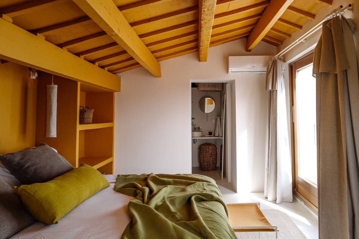 A bedroom with a bed, a wooden ceiling, a window with curtains, and a small table.