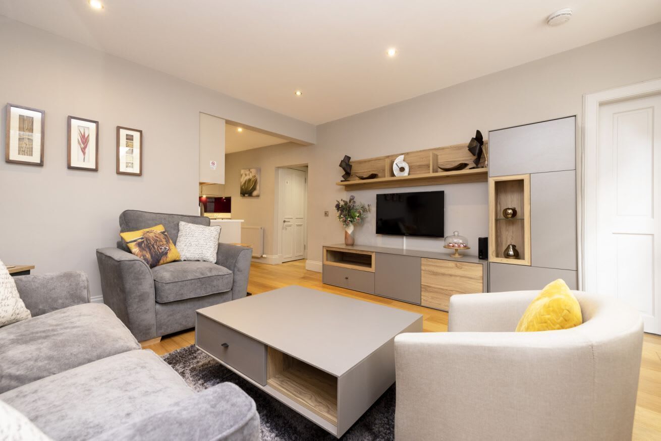 A modern living room with a grey sofa, a white armchair, and a coffee table. There is a flat-screen TV mounted on the wall, a wooden shelf above it, and a door to the right.