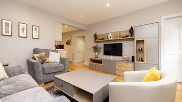 A modern living room with a grey sofa, a white armchair, and a coffee table. There is a flat-screen TV mounted on the wall, a wooden shelf above it, and a door to the right.