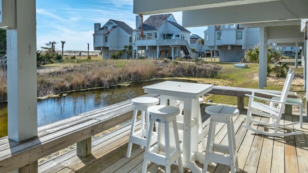 A wooden deck with white barstools and a table, overlooking a body of water and beachfront houses.