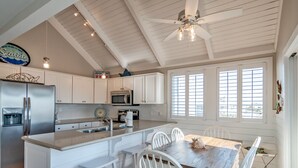 A kitchen with white cabinets, a stainless steel refrigerator, a wooden dining table with white chairs, and a ceiling fan.