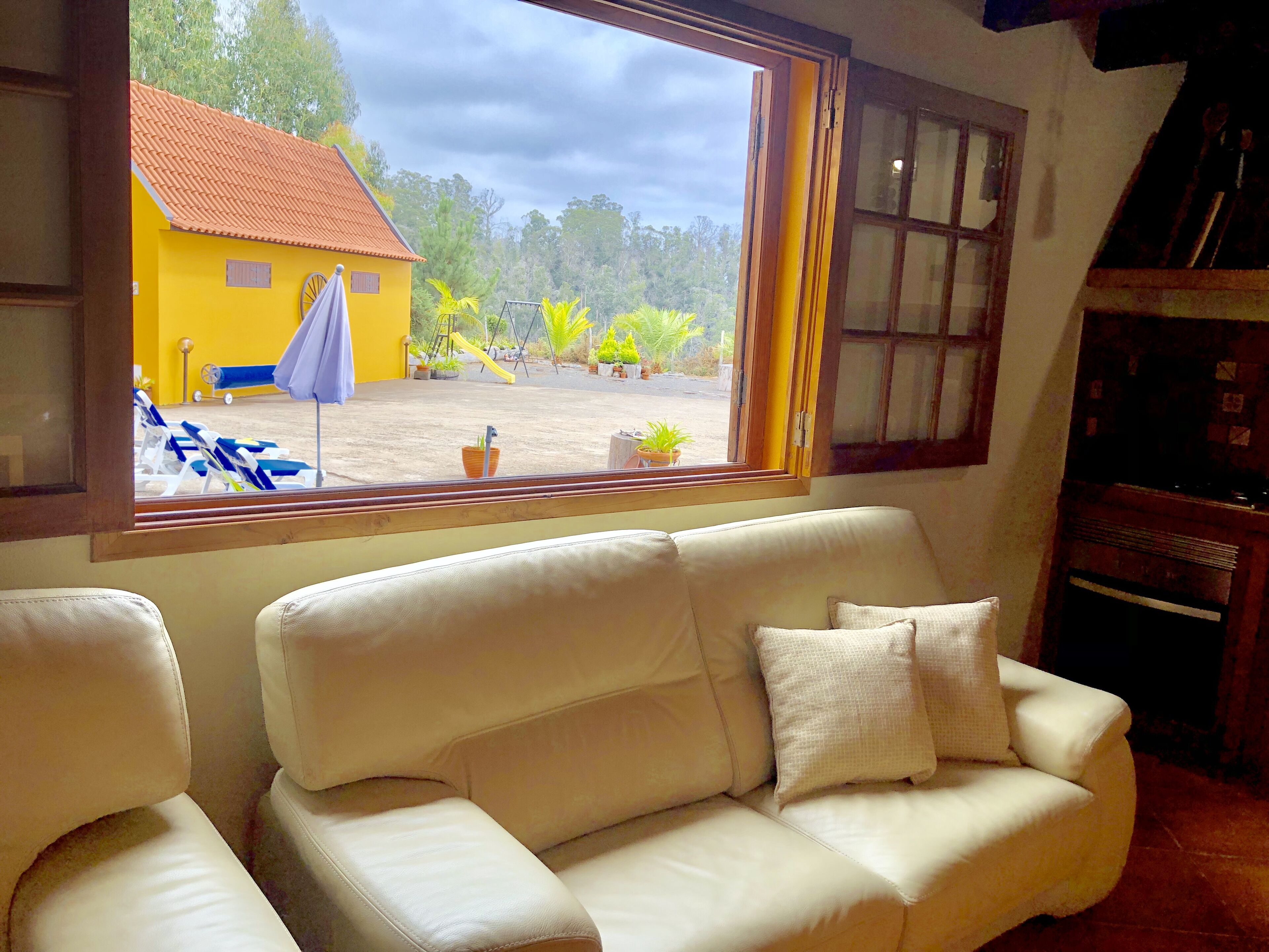 A room with a beige sofa, a window with a view of a yellow building and outdoor furniture, and a wooden cabinet.