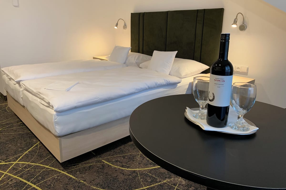 A hotel room with a bed, a bedside table with a bottle of wine and two glasses, and a headboard.