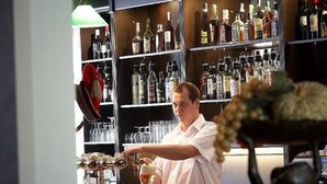 Angolo bar con scaffali pieni di bottiglie di liquori, un barista e un vaso decorativo con fiori.