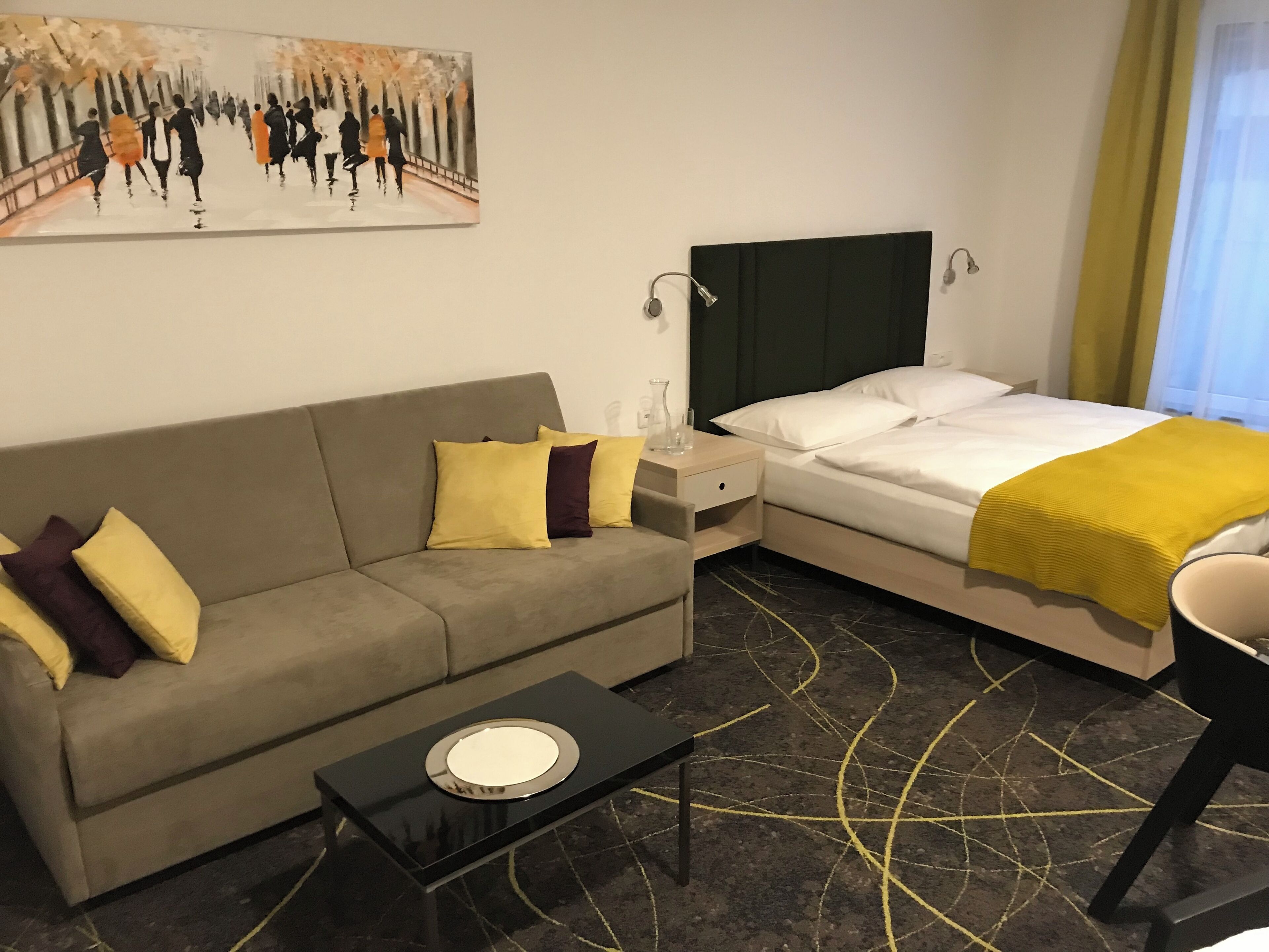 A hotel room with a sofa, a bed with a yellow blanket, a nightstand, and a painting on the wall.