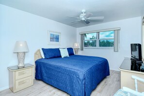 A bedroom with a bed, bedside tables, a ceiling fan, a window with a view, and a desk with a chair.