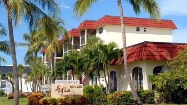 A two-story building with a red roof and white walls, surrounded by palm trees and a well-maintained lawn.