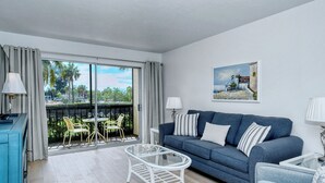 A modern living room with a blue sofa, white armchairs, a glass coffee table, and a balcony with outdoor seating.