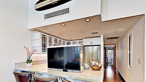 A modern kitchen with a large TV, white cabinets, and a dining area with leather chairs. - Where Sunsets Dazzle Kihei Oceanfront Corner Condo Sweeping Views and Lanai (Kihei)