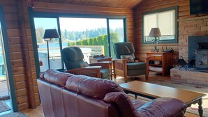 A cozy cabin interior with a leather sofa, two armchairs, a wooden coffee table, a fireplace, and a large window offering a view of the outdoors. - Lake Shoecraft Waterfront Log Home (Stanwood)