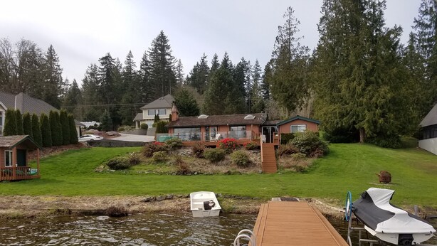 A wooden dock extending into a body of water with a house and greenery in the background. - Lake Shoecraft Waterfront Log Home (Stanwood)