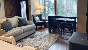 A cozy living room with a sofa, armchair, and a dining table with chairs. A window offers a view of trees. - Panoramic Coeur D'Alene Lake View - 5 Minutes to Town (Coeur D Alene)