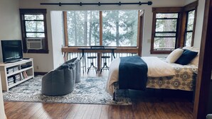 A bedroom with a bed, a television, a desk, and a view of trees through large windows. - Panoramic Coeur D'Alene Lake View - 5 Minutes to Town (Coeur D Alene)