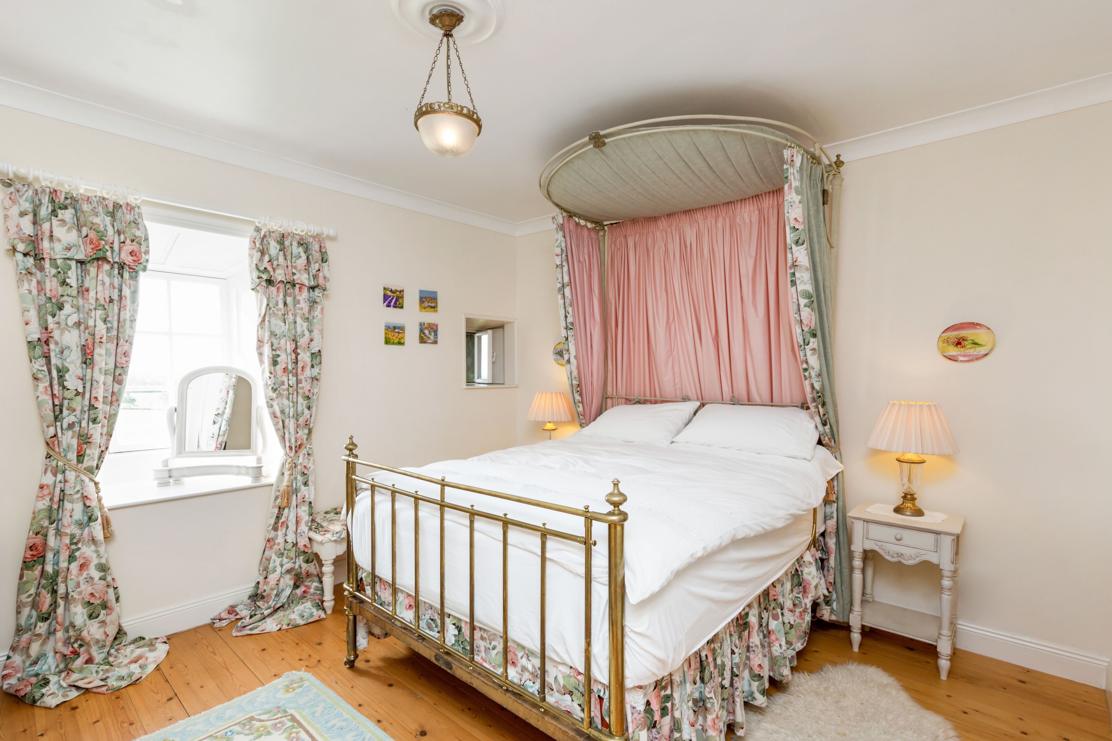 Una cama con dosel adornada con cortinas florales, una mesita de noche con lámpara y una alfombra pequeña sobre piso de madera.