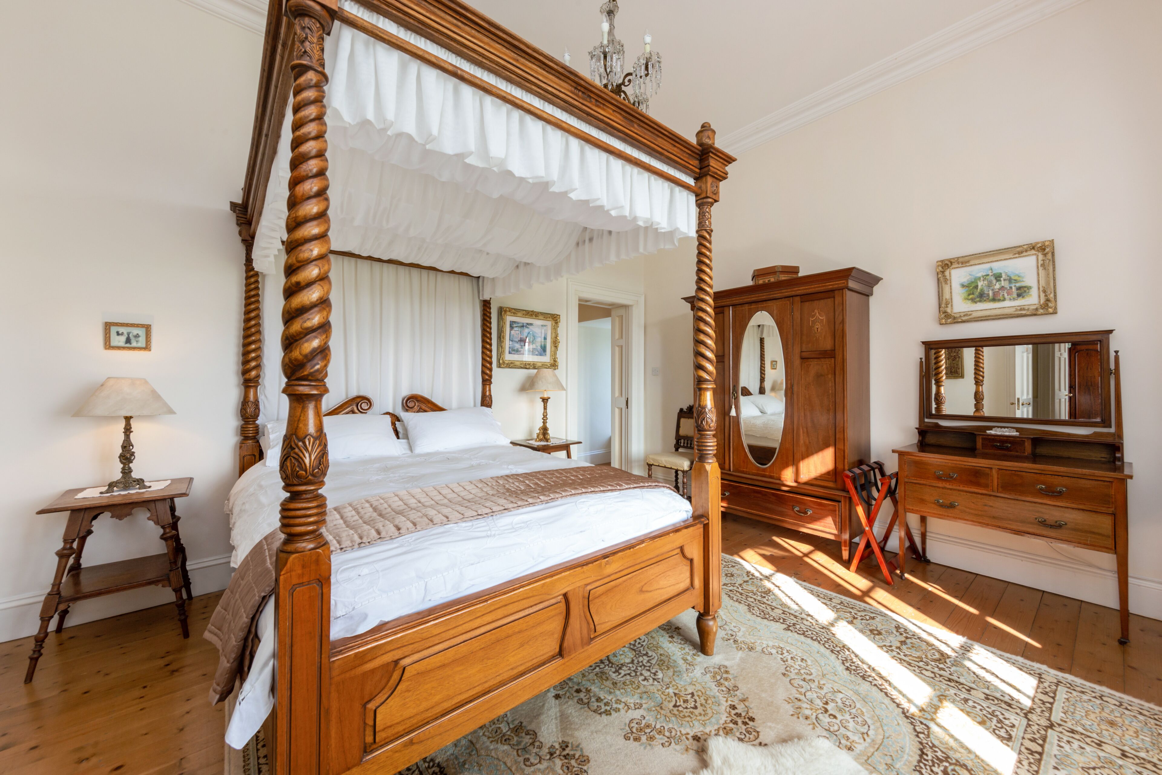 Un lit à baldaquin, une commode en bois, un miroir et un tapis à motifs dans une pièce bien éclairée, dotée d’un plancher en bois.
