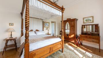 Un lit à baldaquin, une commode en bois, un miroir et un tapis à motifs dans une pièce bien éclairée, dotée d’un plancher en bois.