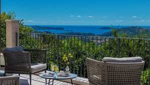Un balcón con muebles de mimbre, una mesa con un florero y vasos, y vistas al mar y las colinas.