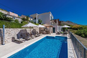 Piscina con sillas de descanso y sombrillas, rodeada de muros de piedra y vista a la montaña.