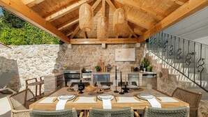 Cocina al aire libre con paredes de piedra, techo de madera y comedor con sillas de mimbre y una mesa de madera para cuatro.