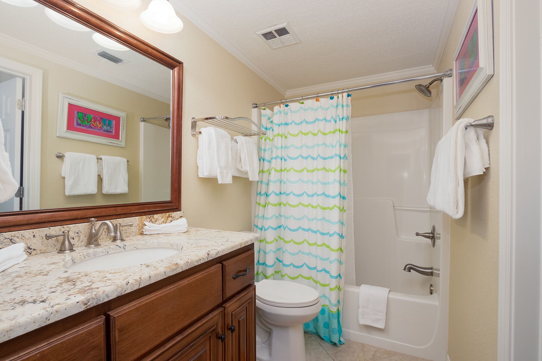 A bathroom with a marble sink, wooden cabinets, a toilet, a shower with a blue and green shower curtain, and a mirror with a picture frame.