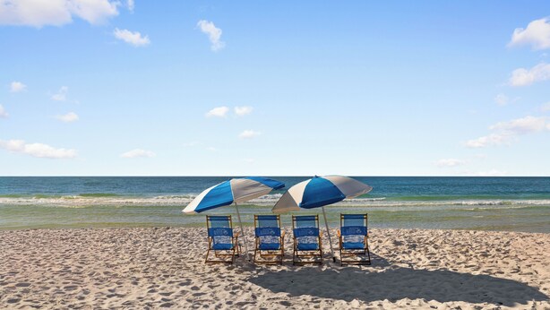 Ein Strand mit vier blauen und weißen Liegen und Sonnenschirmen, einem sandigen Ufer und dem Meer im Hintergrund.