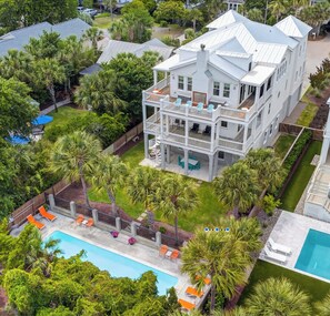 A large two-story house with a pool, surrounded by lush greenery and palm trees.