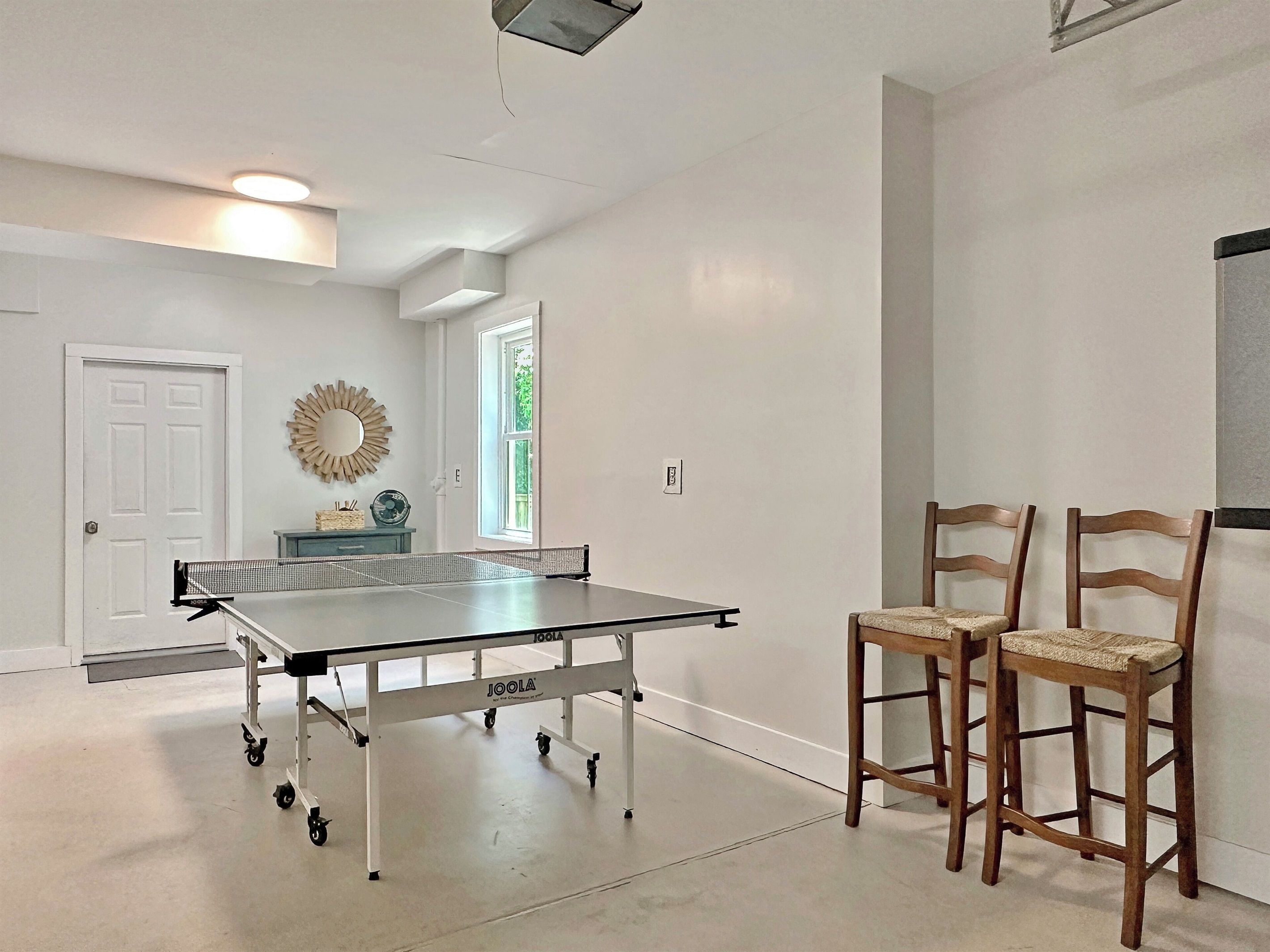 A ping pong table with wheels, two wooden chairs, a white door, and a wall-mounted mirror.