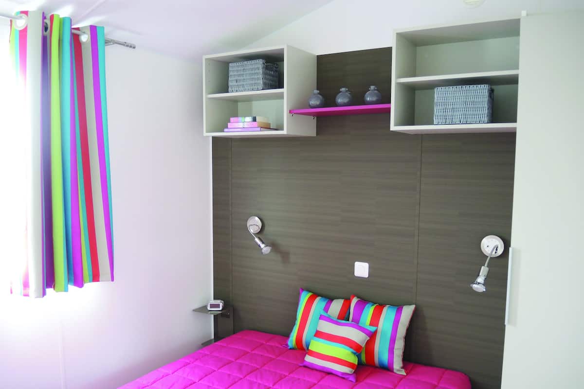 A bedroom with a bed, a nightstand, a shelf with decorative items, and a window with curtains.