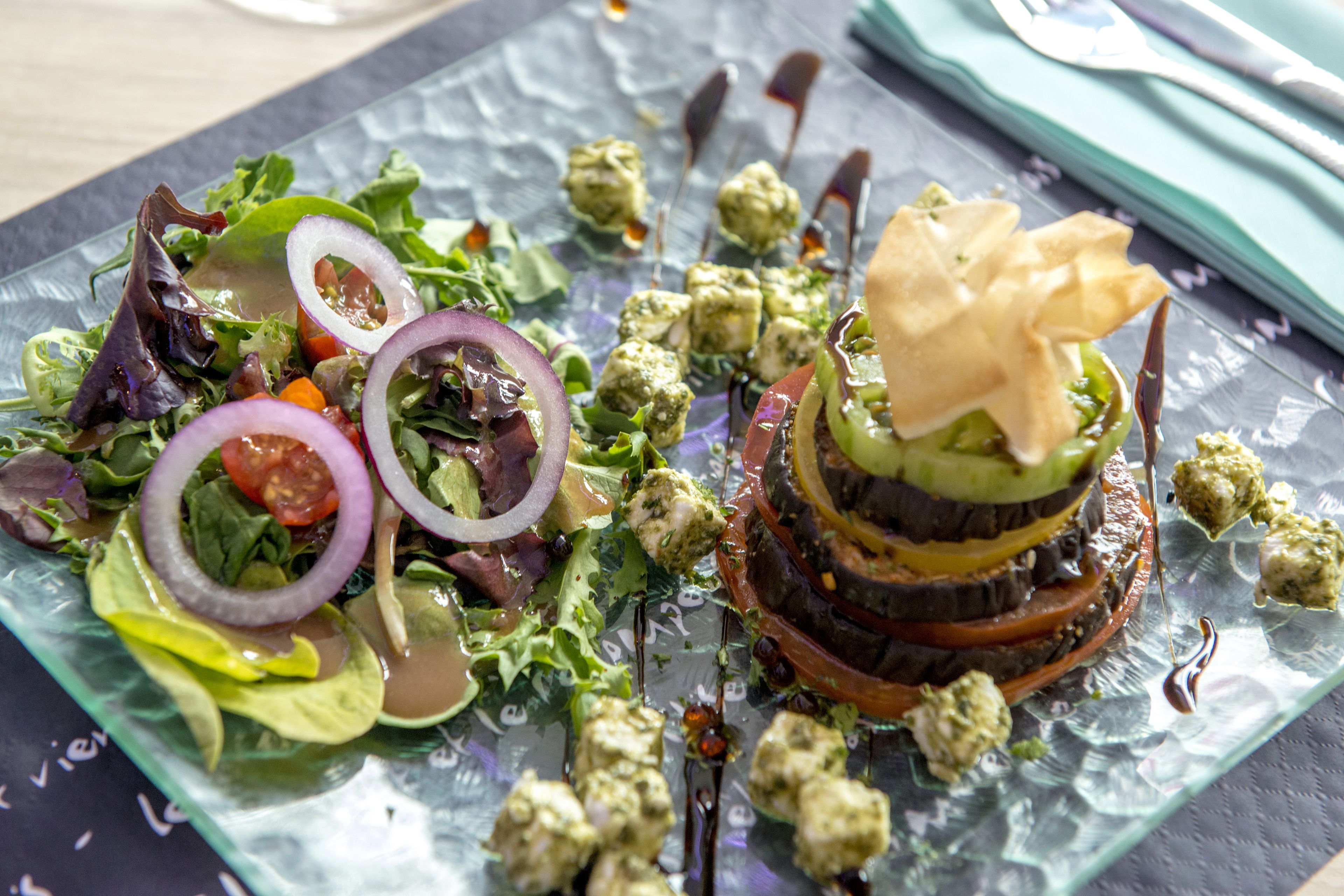 A glass plate with a salad and a layered dish, possibly a burger or sandwich, on a table with a napkin and cutlery.