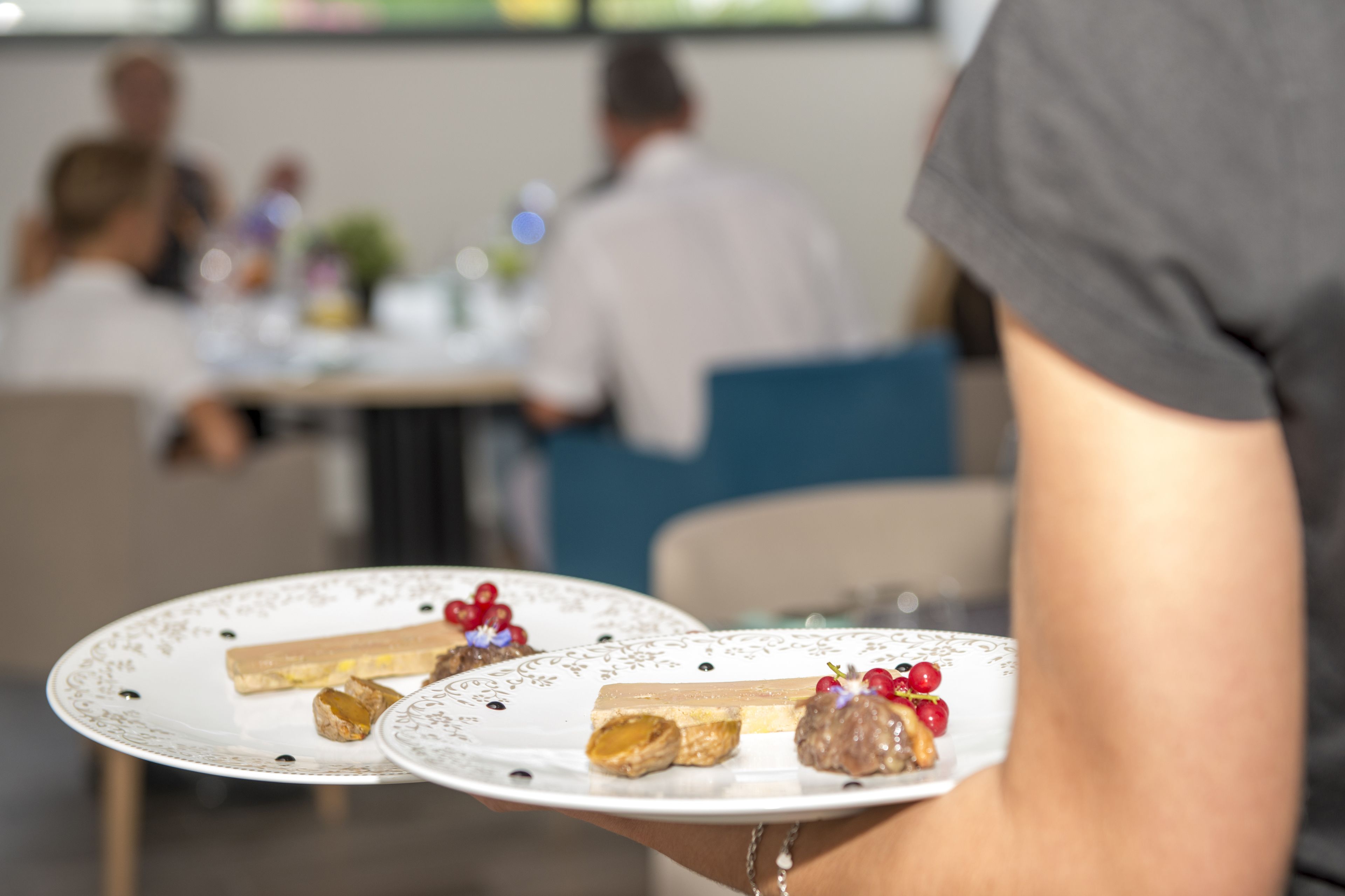 Eine Person hält einen Teller mit Desserts und einer Torte, im Hintergrund verschwommen ein Essbereich.