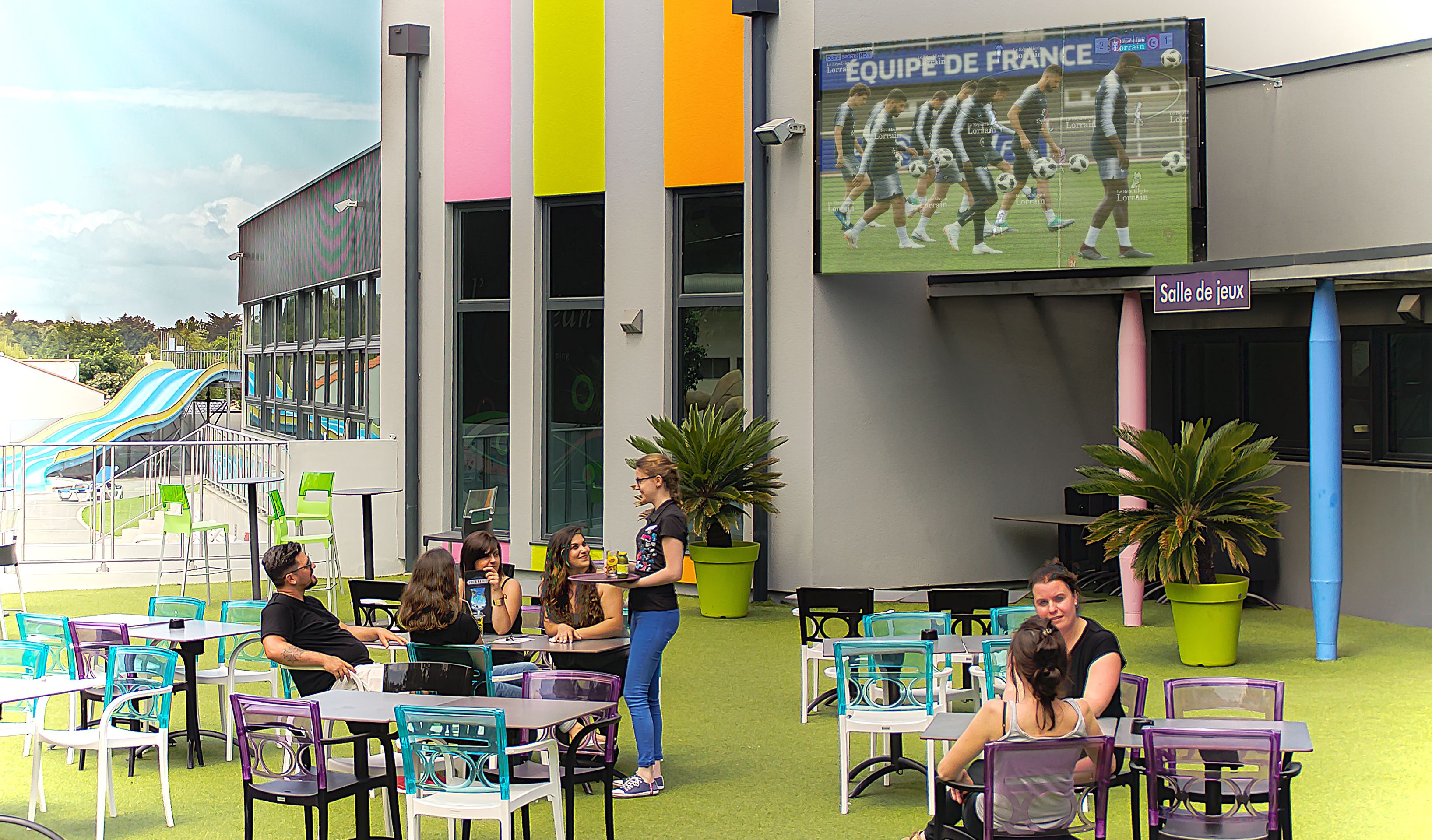 An outdoor seating area with tables and chairs, a building with a large screen displaying a sports team, and a playground with slides.