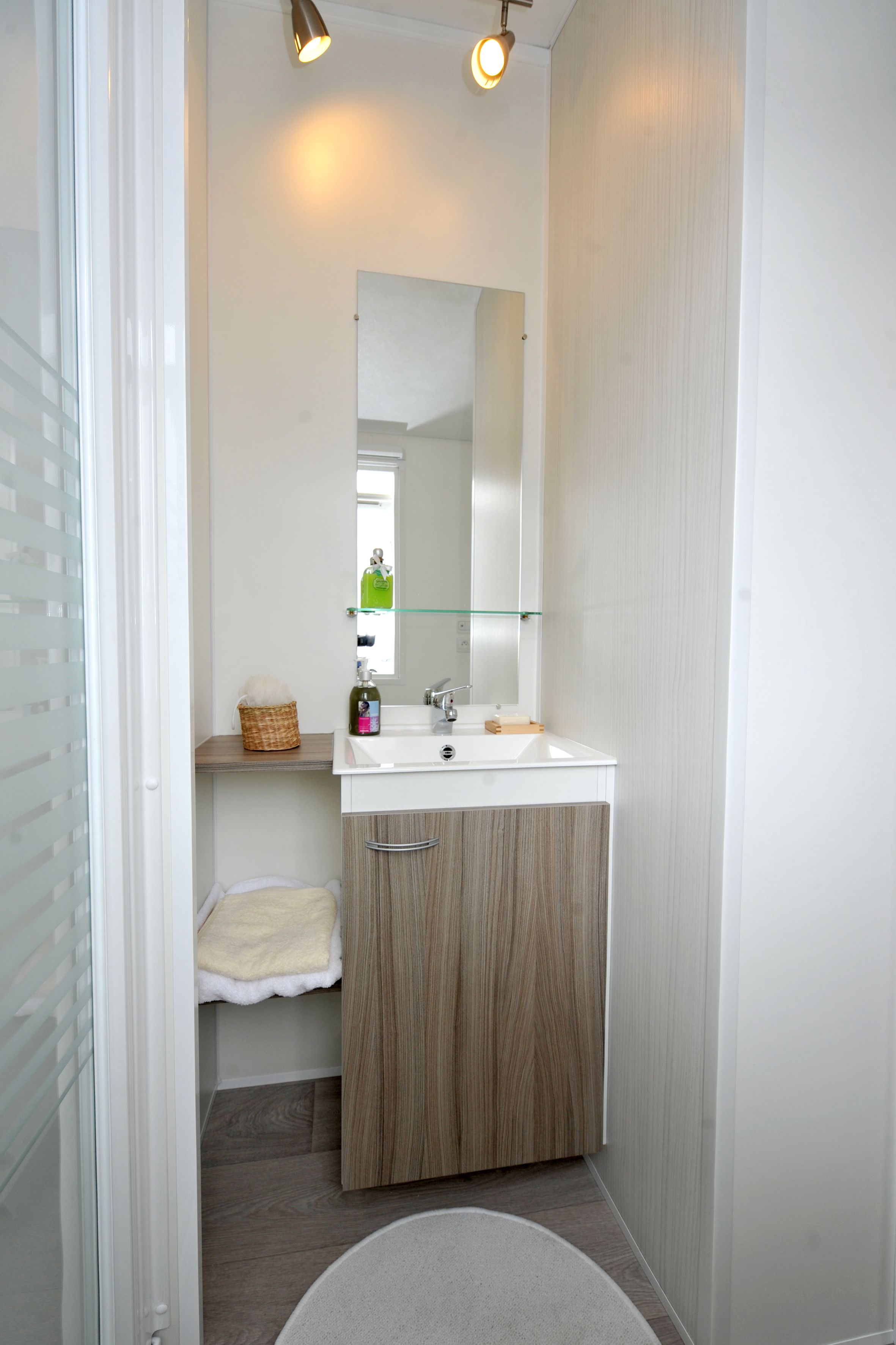 A bathroom with a white sink, wooden cabinet, glass shelf, and a mirror.