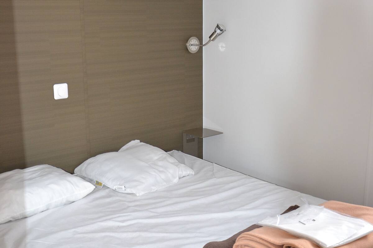 A neatly made bed with white and brown linens, a small shelf above it, and a wall-mounted light fixture.