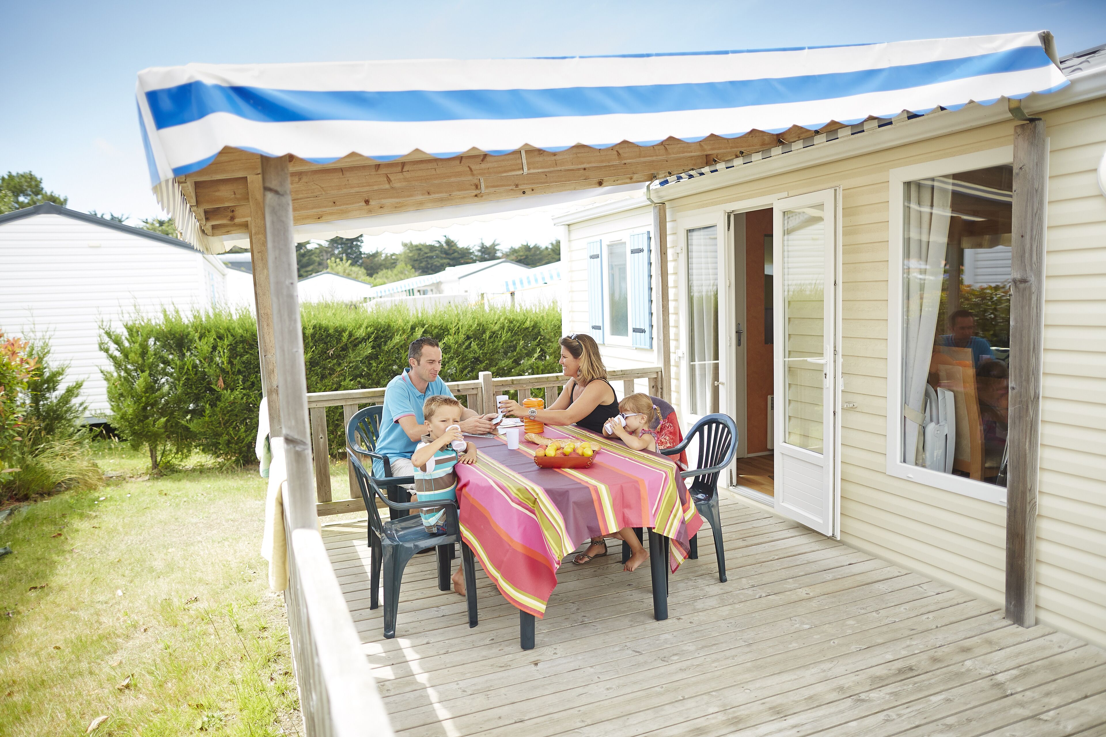 Une famille est assise à une table à l’extérieur, sous une pergola, et la table est recouverte d’une nappe à rayures.