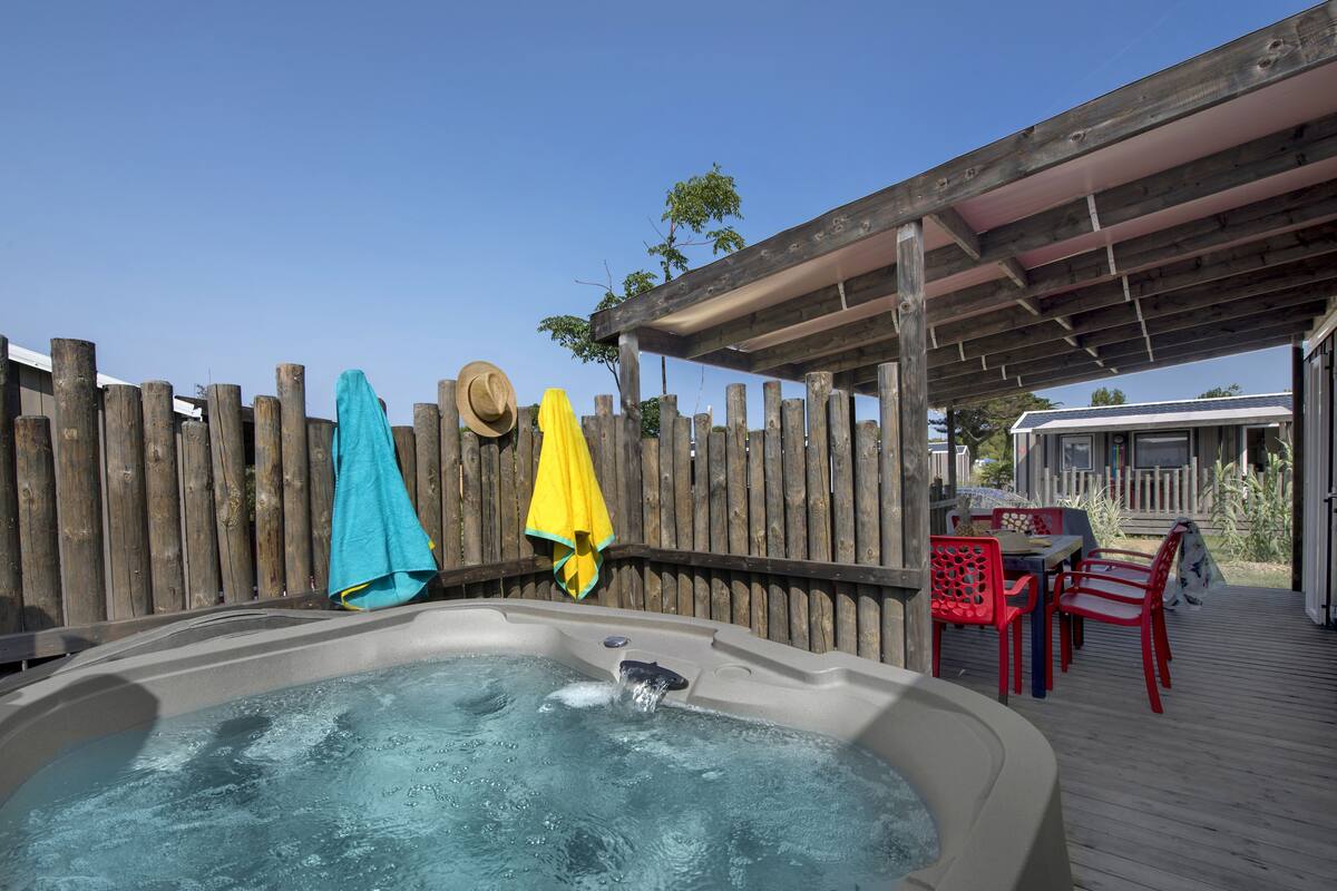 A hot tub with a wooden deck and red chairs, a wooden fence, and a covered area with a table and chairs.