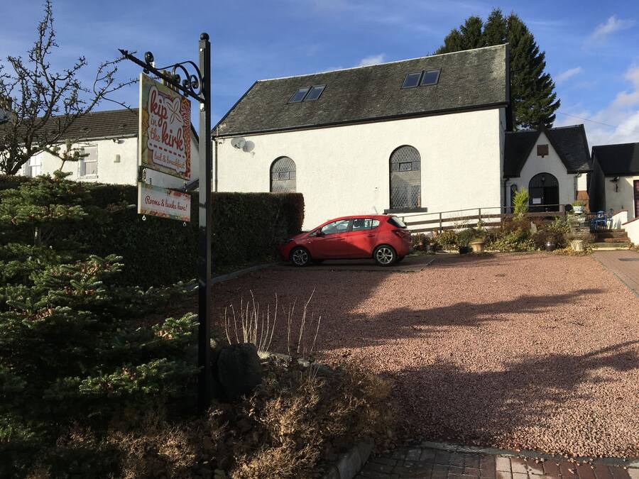 A red car parked in front of a white building with a gravel driveway and a hedge.
