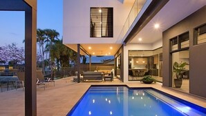Ein modernes Haus mit Swimmingpool, Sitzgelegenheiten im Freien und einer durch große Fenster einsehbaren Küche.