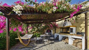 An outdoor patio with a pergola covered in flowering plants, a stone-tiled floor, and a barbecue grill. - VILLA DEUS (Santorini,Thira)