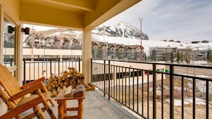 Ein Balkon mit Holzkästen und einem Tisch, ein Holzstoß und Blick auf einen verschneiten Berg sowie Gebäude.