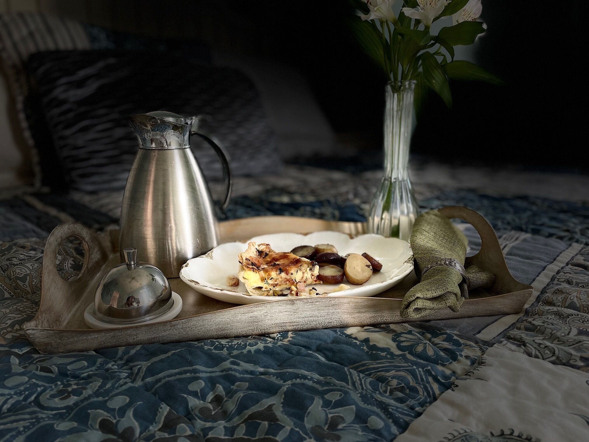 A tray with a plate of food, a coffee pot, and a vase with flowers on a patterned bedspread.